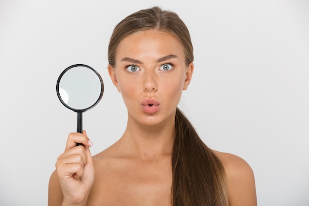 Beauty portrait of healthy shirtless woman with long brown hair holding magnifying glass, isolated