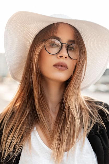 Beauty portrait lovely young woman with clean tender skin with brown hair with sexy lips in fashion glasses in white top in straw hat in youth black blazer in sunny bright day outdoors. Elegant lady.