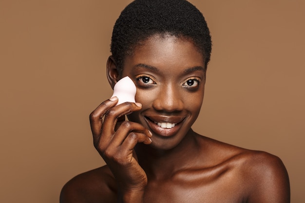 Beauty portrait of pretty young half-naked african woman holding makeup puff isolated on brown