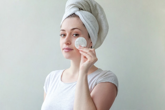 Beauty portrait of smiling woman in towel on head with soft healthy skin removing make up with cotton pad isolated on white background.