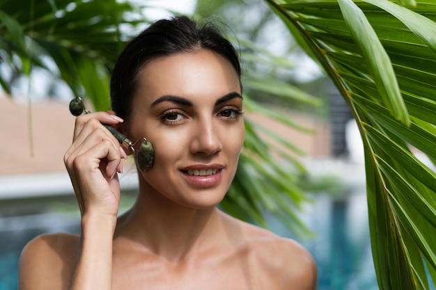 Beauty portrait soft skin Female model with natural makeup and healthy skin behind a green leafy plant Portrait of a beautiful girl with nude nails bare shoulders holding roller massager outdoor