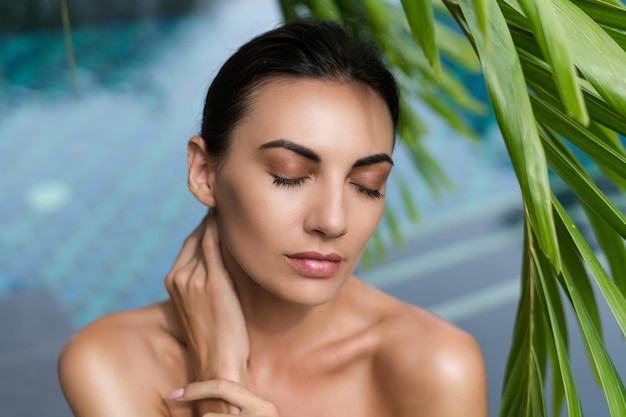 Beauty portrait soft skin Female model with natural makeup and healthy skin behind a green leafy plant Portrait of a beautiful girl with nude nails bare shoulders