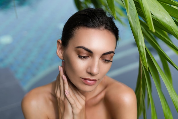 Beauty portrait soft skin Female model with natural makeup and healthy skin behind a green leafy plant Portrait of a beautiful girl with nude nails bare shoulders