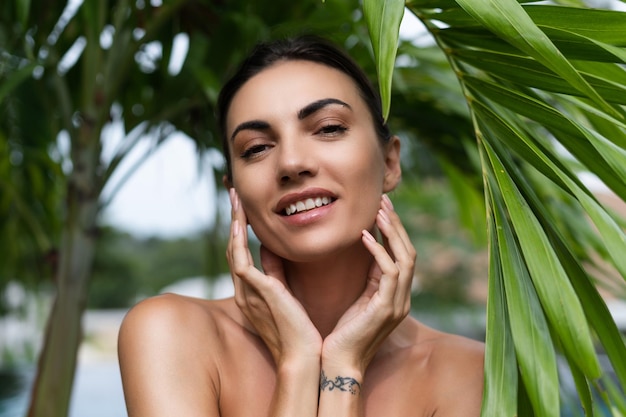 Beauty portrait soft skin Female model with natural makeup and healthy skin behind a green leafy plant Portrait of a beautiful girl with nude nails bare shoulders