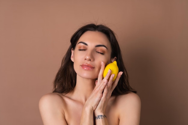 Beauty portrait of topless woman with perfect skin and natural makeup on a beige background holds citrus lemon vitamins c for skin