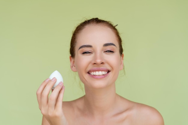 Beauty portrait of young topless red hair woman with bare shoulders on green background with makeup foundation sponge