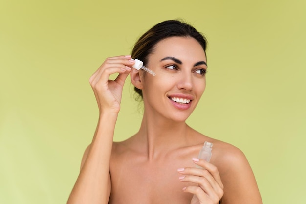 Beauty portrait of young topless woman with bare shoulders on green background with perfect skin and natural makeup holds serum for youth and skin hydration