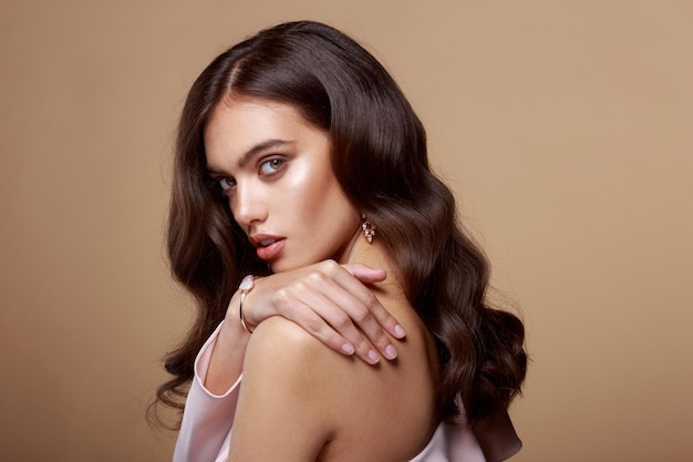 Beauty portrait of a young woman with bare shoulders, brown wavy hair, model with bracelet and earrings, isolated beige background.