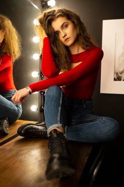 Beauty portrait of a young woman with curly hair sitting on wooden table posing near mirror looking at camera