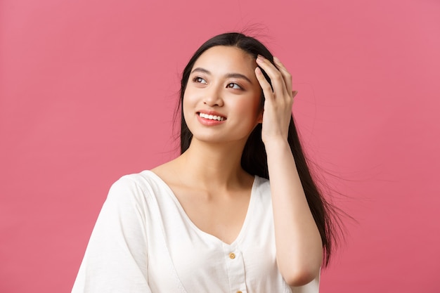 Beauty products advertisement, haircare and women fashion concept. Sensual romantic asian woman looking away with dreamy smile while running hand in hair, standing pink background happy.