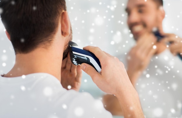 beauty, shaving, grooming and people concept - close up of young man looking to mirror and shaving beard with trimmer or electric shaver at home bathroom over snow