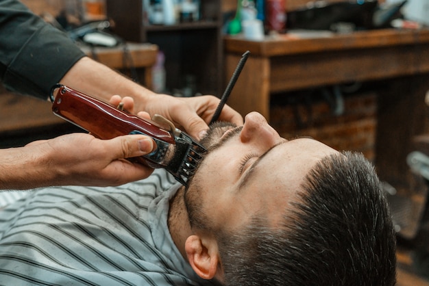 Beauty shop for men. Shaving a beard in a barbershop. Barber cuts his beard with a razor and clipper. close up Brutal haircuts. Hairdresser equipment. Selective focus.