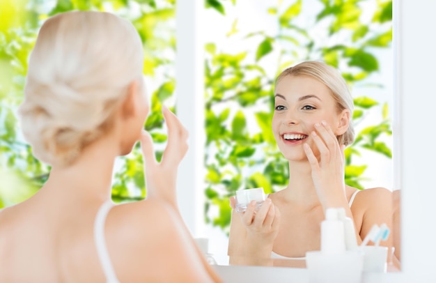 beauty, skin care and people concept - smiling young woman applying cream to face and looking to mirror at home bathroom over green natural background