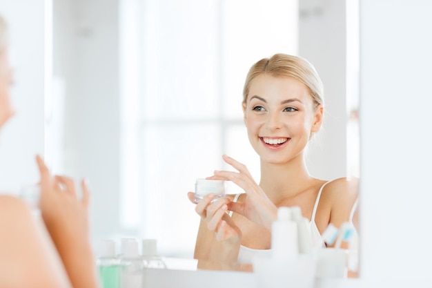 beauty, skin care and people concept - smiling young woman applying cream to face and looking to mirror at home bathroom