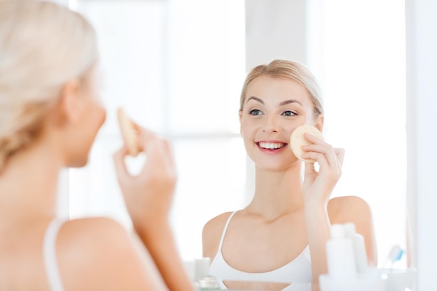 beauty, skin care and people concept - smiling young woman washing her face with facial cleansing sponge at bathroom