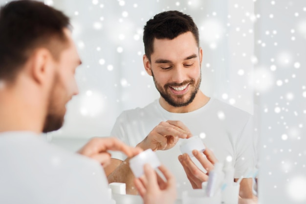 beauty, skin care, winter and people concept - smiling young man applying cream to face and looking to mirror at home bathroom over snow