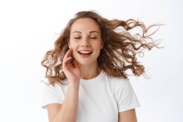 Beauty and skin care Young blond woman touching soft clean face and close eyes enjoying soft wind blowing at her hairstyle hair flying in air standing over white background