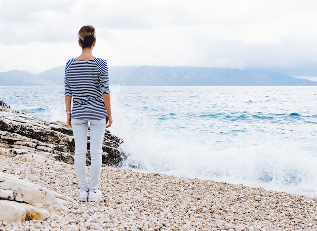 Beauty stylish young woman enjoys standing on pebble stone shore at the seaside in cloudy summer day and looking away. 