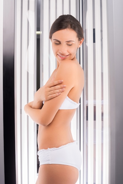 Beauty sunbathing. Side view of attractive young woman standing in tanning booth and keeping eyes closed