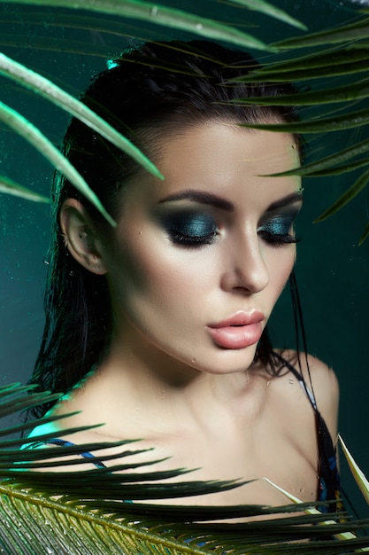 Beauty woman in palm leaves wet makeup, tropical portrait girl in green swimsuit in branches palm tree in studio, smoke and raindrops on glass. Sexy woman with bright green makeup