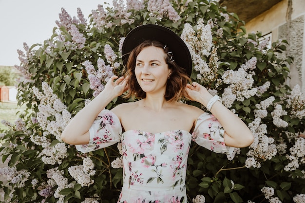 Beauty woman in a romantic light dress with bare shoulders and a hat in a spring garden with blooming lilacs