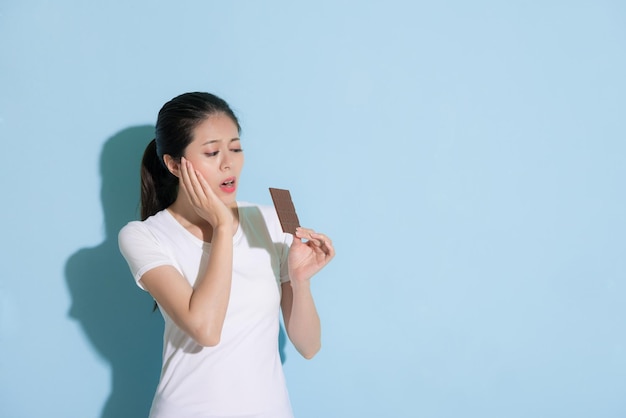 beauty young woman looking at chocolate cookie standing on blue wall background and feeling teeth painful because she having tooth decay problem.