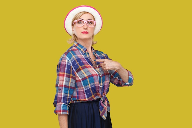 Because I am. Portrait of proud serious modern stylish mature woman in casual style with hat and eyeglasses standing, pointing herself and looking at camera. studio shot isolated on yellow background.