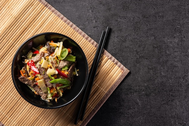 Beef, vegetables and sesame seeds in black bowl on black slate background