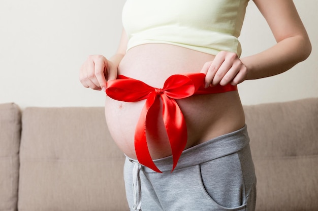 Belly of a pregnant woman tied with a red bow on a home background