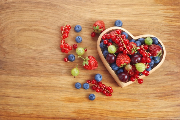 Berries mix in in a heart shaped bowl spilled on rustic wooden table