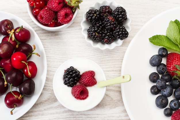 Berries in white bowls and plates on a white wooden table in a top view