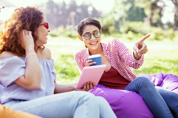 Best day. Cheerful curly-haired woman holding a tablet and talking with her friend