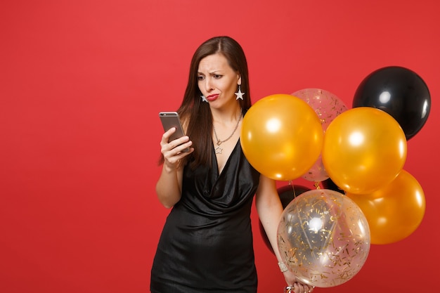 Bewildered young woman in little black dress holding air balloons using mobile phone while celebrating isolated on red background. Valentine's Day Happy New Year birthday mockup holiday party concept.