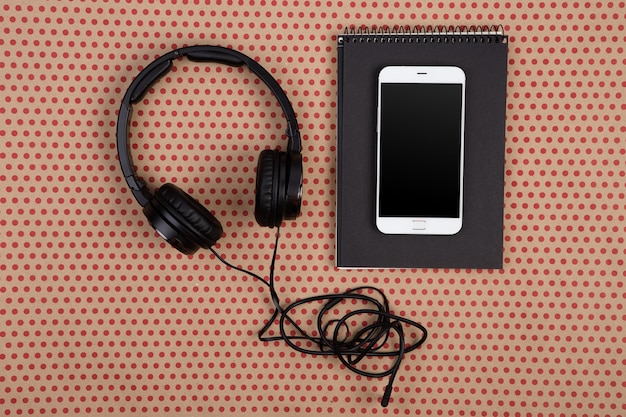 Photo big black headphones white smartphone and black notepad on craft paper background in red polka dots