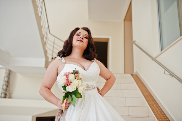 Big breasts brunette bride with wedding bouquet posed at wedding hall on stairs