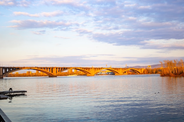 Big bridge in the rays of the setting sun across the river. Cloudy sky. Communal bridge, Yenisei river, Krasnoyarsk, Siberia, Russia.