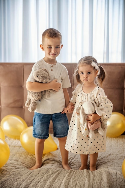 Big brother and his sister are standing on a bed surrounded by balloons and hugging