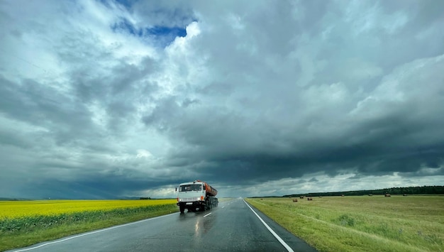 Big car moving across the wet road after rain. Yellow field and cloudy sky around. Travel by car con