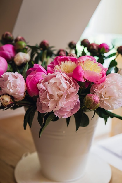 Big fresh pink and white peony blooming bouquet in a white metal bucket on a wooden table in a light bright modern beautiful apartment Spring home decor or present idea Vivid summer colors