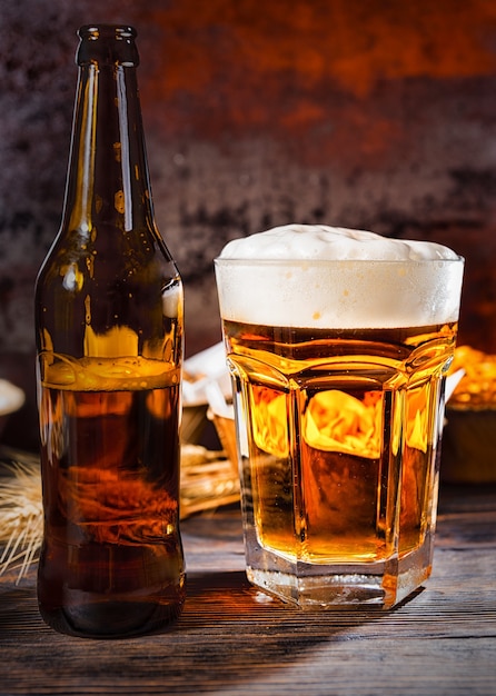 Big glass with freshly poured beer and head of foam near bottle and plates with snacks on dark wooden desk. Food and beverages concept