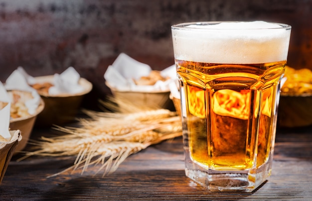 Big glass with freshly poured beer near plates with pistachios, small pretzels and peanuts on dark wooden desk. Food and beverages concept