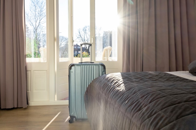 Big green suitcase stays in the room in hotel near window or balcony with sunlight