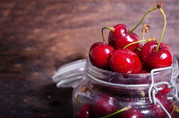 Photo big red cherries in a glass jar on dark wooden background with copy space.