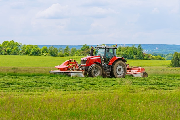 A big red tractor with two mowers mows the green grass on a silo.