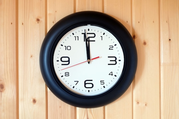 Big round wall clock with a black rim with arrows showing twelve o'clock hangs on brown wooden wall from vertical planks horizontal view indoor close up