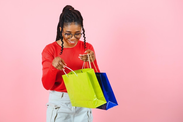 Big Sale Excited afro girl with shopping bags touching sunglasses looking at camera over pink studio wall