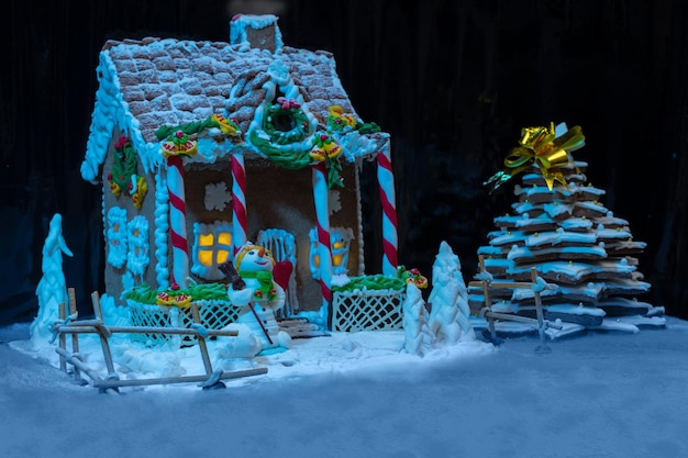 Big snow-covered homemade gingerbread house with lights inside, gingerbread Christmas tree and a sugar mastic snowman at night. Mockup for seasonal offers and holiday post card