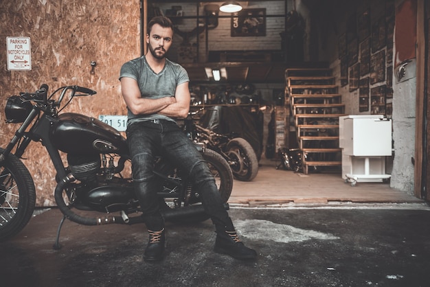 Bike is my life. Handsome young man keeping arms crossed and looking at camera while leaning at his bike with repair shop in the background