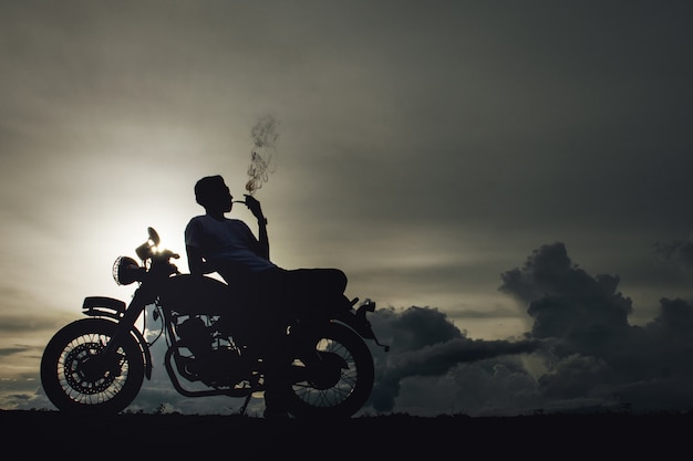 Biker man sitting smokes with his motorbike beside the natural lake and beautiful.