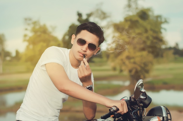 Biker man sitting smokes with his motorbike beside the natural lake and beautiful.